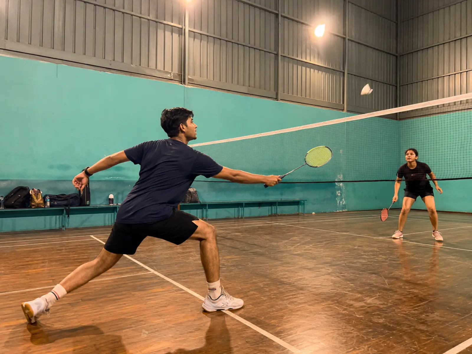 Two young players on an indoor badminton court in India mid-rally under bright court lights, rackets raised, warm candid phone-photo energy
