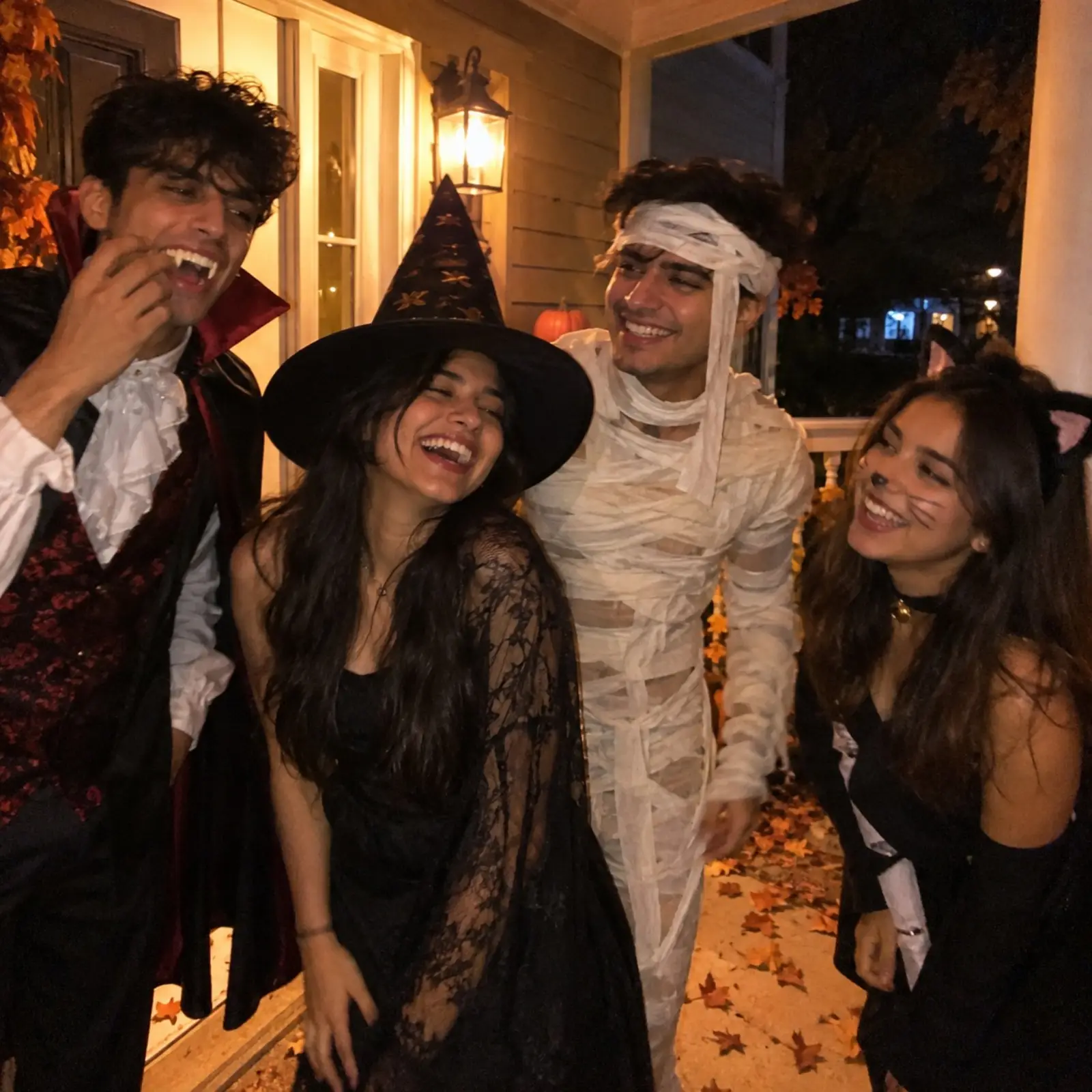 Friends in homemade Halloween costumes laughing outside a lit doorway at night, warm porch light and autumn leaves