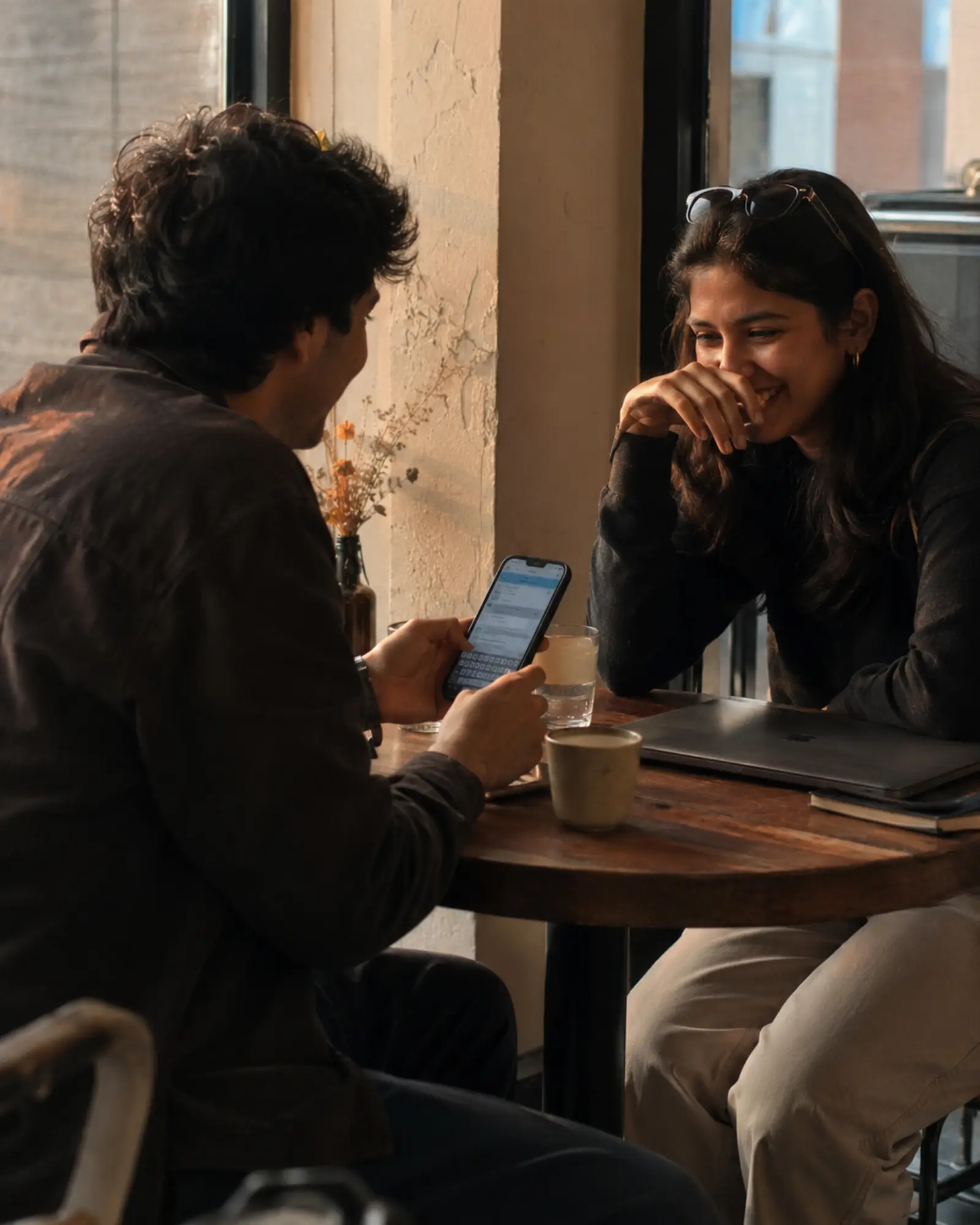 Two coworkers at a cafe table, one showing their phone with a casual text draft, warm afternoon light, laptops closed to the side