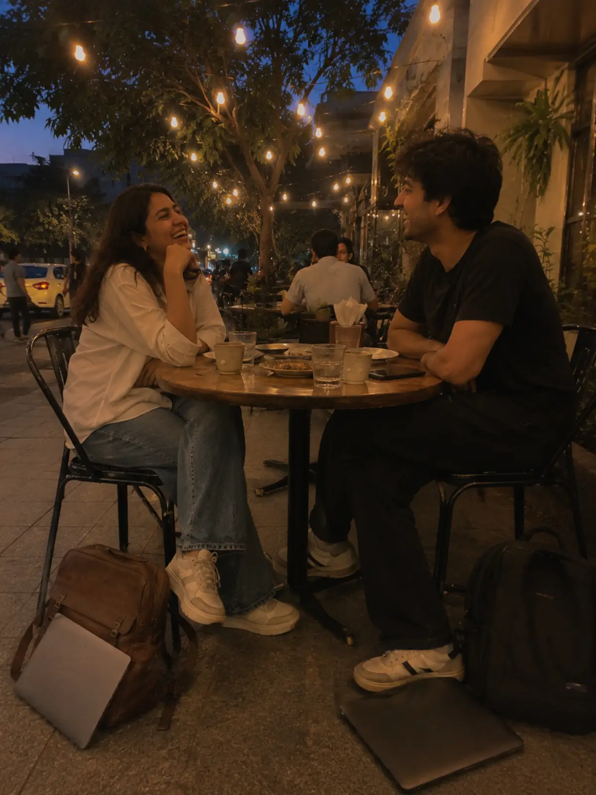 Two friends laughing at an outdoor cafe table after work, laptops on the ground beside them