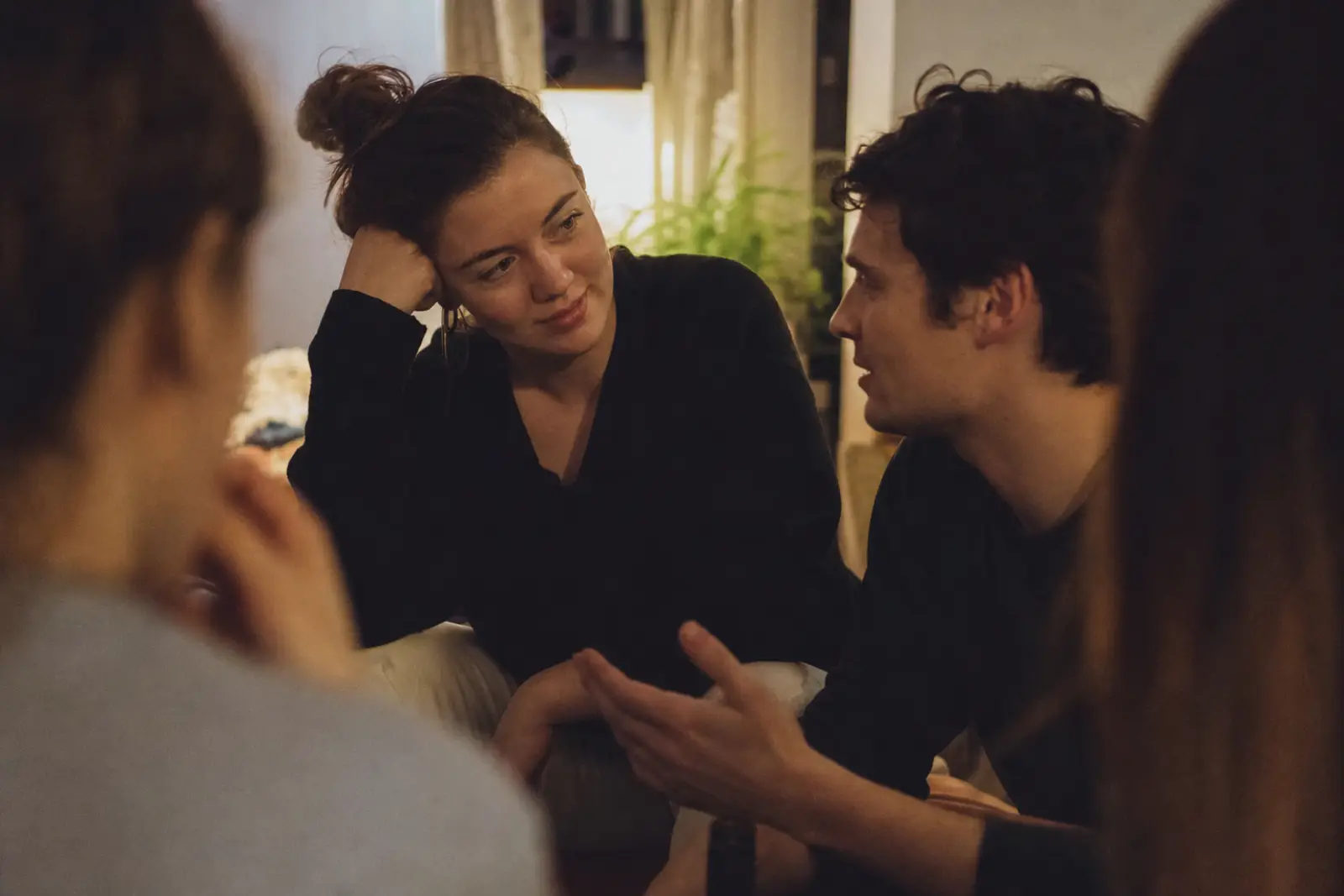 A crowded house party living room at night, two people mid-conversation near a window while the rest of the room blurs into soft motion
