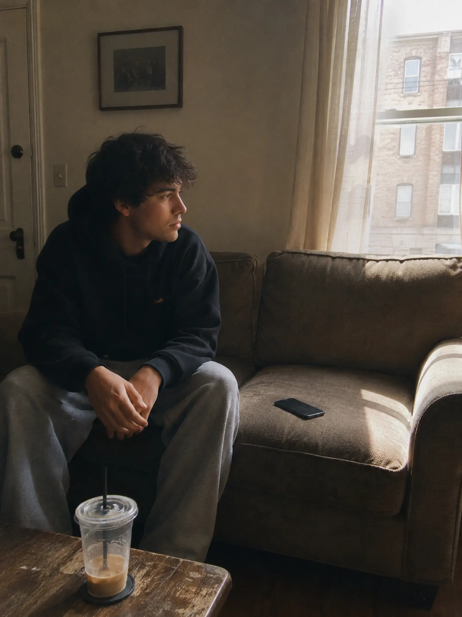 Young graduate sitting alone on a Saturday afternoon in a quiet apartment, phone face-down, sunlight on an empty couch that used to hold friends