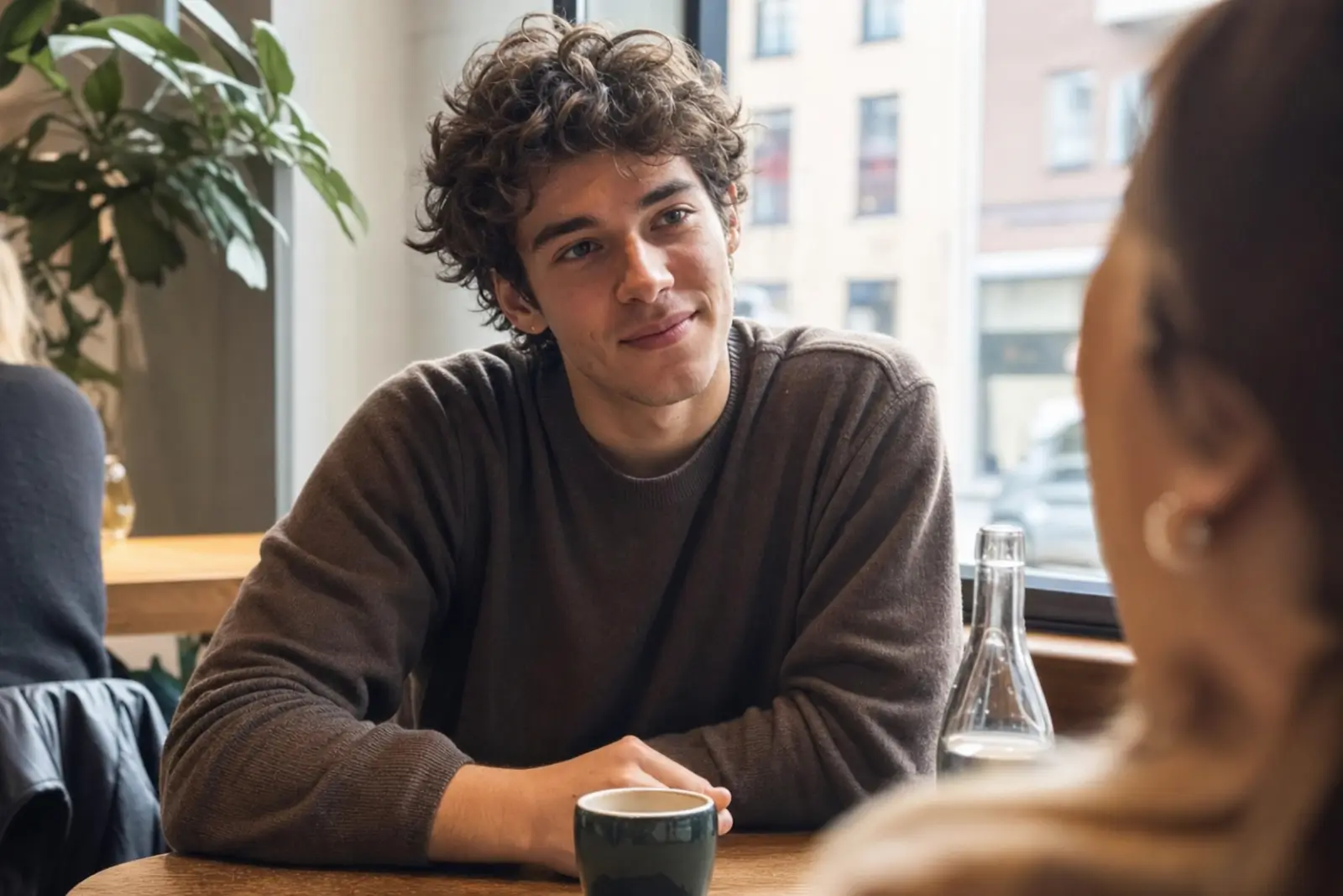 Two young friends talking across a café table, one leaning in mid-sentence while the other listens, phones face-down, warm afternoon light